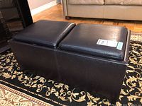Side angle view of black faux leather ottoman with cushioned tops, showing overall shape and material.