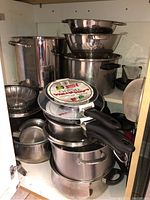 View of assorted stainless steel pots, large soup pots, colanders, and smaller sauce pans stacked inside a kitchen cabinet.