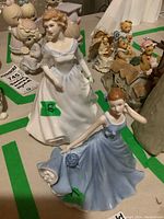 Two Royal Doulton porcelain figurines marked with green tape and auction labels on a table showing detailed views of the figurines