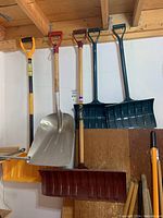 Photo showing five snow shovels hanging on a wall including yellow-handled, metal blade, red plastic, and two green plastic shovels