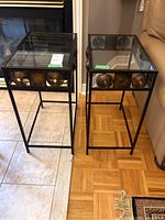 Front view of two rectangular side tables with black metal frames and glass tops, showing decorative circular metal accents on upper sides.