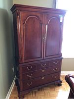 Front view of Thomasville wooden armoire dresser cabinet with two paneled doors above three drawers, brass handles, standing on four carved feet.
