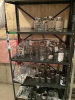 Black metal utility shelf unit with four shelves filled with various glass jars and containers. Photo shows full unit in a basement setting.