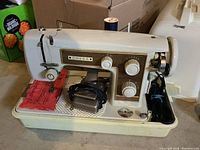 Side view of vintage Omega sewing machine with attached foot pedal, spool of thread, red fabric under needle, and carrying case visible.