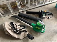 Two yard power tools on concrete floor: Greenwood electric chainsaw with blade cover and Weed Eater 2540 electric leaf blower with collection bag attached.