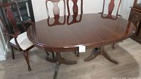 Oval cherry finish dining table on dual pedestal bases with some surface scratches, surrounded by chairs.