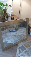 Mirror leaning against a rug and table, showing clear reflective surface and a thick wood-like frame.