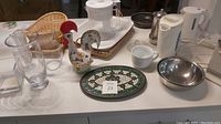 Overview of various kitchen items including a ceramic rooster figurine, glassware, serving plate, kettles, metal bowl, and other kitchen utensils arranged on a counter.