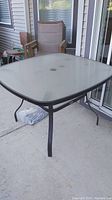 Square glass top patio table with black metal frame and umbrella hole visible in center. Shows table surface and leg structure.