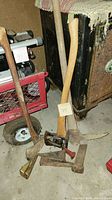 Various axes with wooden handles and several heavy metal wedges, including a specialized wedge with red handle, placed on a concrete floor near old metal cabinet and red hand cart.