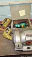The lot on a table showing the bench grinder, yellow drill, green hand grinder in wooden box, and drill bit tools.