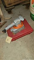 Electric palm sander by Black and Decker shown on a red plastic case in a workshop or storage area.