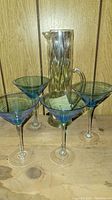 Four blue stem martini glasses and one clear glass pitcher on wooden surface, possibly with chips and cracks.