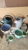 Photo showing green and cream McCoy ceramic planters, a black metal stand, two watering cans (green and galvanized metal), and a small green ceramic planter on the ground.