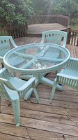 Photo showing full round glass top table with four turquoise plastic chairs on wooden deck, set outdoors with trees in background.