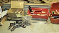 Gray metal toolbox open showing clamps, plastic bag with hardware parts, and small hand tools