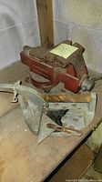 Photo of a red metal vise grip and a gray metal miter box saw on a wooden surface against a wall, showing wear and rust.