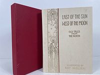 Photo of the book upright with soiled maroon slipcase to the side, showing the cream cover with decorative title and illustration elements.