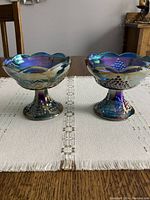 Two matching carnival glass candlesticks side by side on a white table runner with detailed grape and leaf motifs visible and iridescent blue-purple colors.