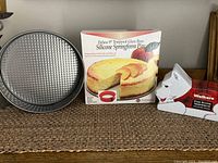 Photo depicts two metal springform pans, one 10-inch and one 9-inch with silicone ring, the 9-inch one in box. Also a Walker's Pure Butter shortbread Westie dog shaped tin.