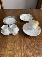 Photo of dual egg cups in white holder, white cup and saucer with floral design, owl salt and pepper shakers, and two decorative saucers arranged on wooden surface.