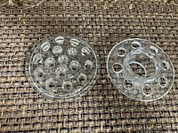 Two clear glass flower frogs side by side on woven fabric surface. One is solid with small holes, other has open center and larger holes.
