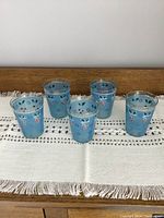 Front view of five blue antique lemonade glasses with hand painted flowers arranged in a row on a white textured cloth runner on a wooden surface.