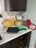 Photo of kitchen lot items on countertop showing Tupperware, bamboo steamer, griddle pan, glass jar, and red silicone kitchen tools