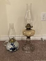 Photo showing two oil lamps, one with a ceramic floral base, the other tall with clear glass components and metal burner.