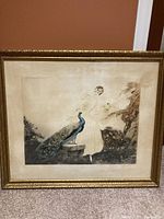 Framed picture showing woman in white dress with peacock perched on a stone pedestal, in an outdoor setting with trees. Signed artwork in decorative gold-tone frame with some aging and discoloration visible.