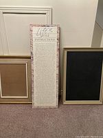 Wide image showing the Life's Little Instructions framed poster board standing between two other framed items, one blackboard and one framed panel