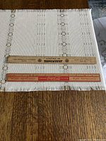 Photo shows two old wooden rulers lying on a white and beige woven cloth. Top ruler is branded Supertest with measurements and black text. Bottom ruler is Coca Cola branded with red advertising text.