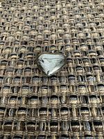Close-up of the silver heart pendant resting on woven fabric, showing shape and surface details.