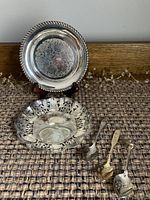 Lot of silver plated items including a butter server, two decorative dishes (one ornate cut work and one embossed), and three souvenir spoons photographed on a textured surface in front of wooden backdrop.