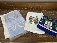 Four tea towels arranged showing new Harry & Megan towel in package, white cotton towel with floral print, and two blue-themed towels with Dylan Thomas and bard portraits.