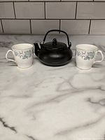 Photo showing two Royal Albert More Silver Maple mugs flanking a black cast iron teapot on a marble countertop with a tiled backsplash.