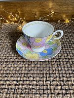 Cup and saucer set with colorful floral pattern and gold accents on a woven brown surface with decorative lights.