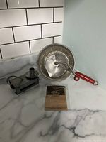 Photo of Foley food mill, metal matchbox holder, and original paper packaging/manual on countertop.