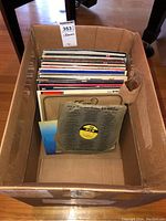 Photo showing the cardboard box with multiple vinyl record albums, visible spine and edges, some wear showing.