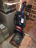 Full view of Bissell carpet cleaner standing upright showing power cord and body in used condition in basement setting.