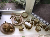 Full set of Denby stoneware dishes spread on white surface near window with outdoor view, showing all the items together.