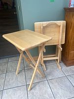 Three wooden folding TV tables with light natural wood finish, one table open and two folded next to the wooden stand.