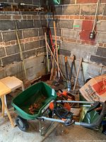 View of the corner with various garden tools including rakes, broom, and the green Scotts Turf Builder lawn spreader with orange adjustment dial sitting on the ground.