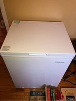 Full front–left angle view of white chest freezer on hardwood floor with books in foreground