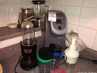 Kitchen counter showing Magic Bullet blender with jars, Oster blender with glass container, Keurig coffee maker, and small white electric coffee grinder with power cord coiled.