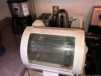 Photo showing Baby George roaster in front with a visible heating element inside, toaster, electric kettle, white cordless kettle in back, and coffee maker partially visible.