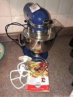 Blue KitchenAid Artisan stand mixer with stainless steel mixing bowl, plastic splash guard, and several attachments including a flat beater and wire whisk along with instruction manual visible.