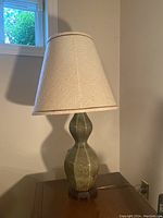 Full view of the lamp on a wooden table showing beige fabric shade and sculpted ceramic body in muted green.