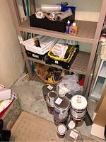 Shelf with various paint cans stacked on the floor below and containers and painting supplies on shelves above.