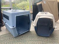 Two resin pet carriers side by side: a blue and gray Vari-Kennel on left, and a light gray Petmate Kennel Cab with blue front door on right. Each has a metal wire door with latch. Seen indoors on tile flooring with other items in background.